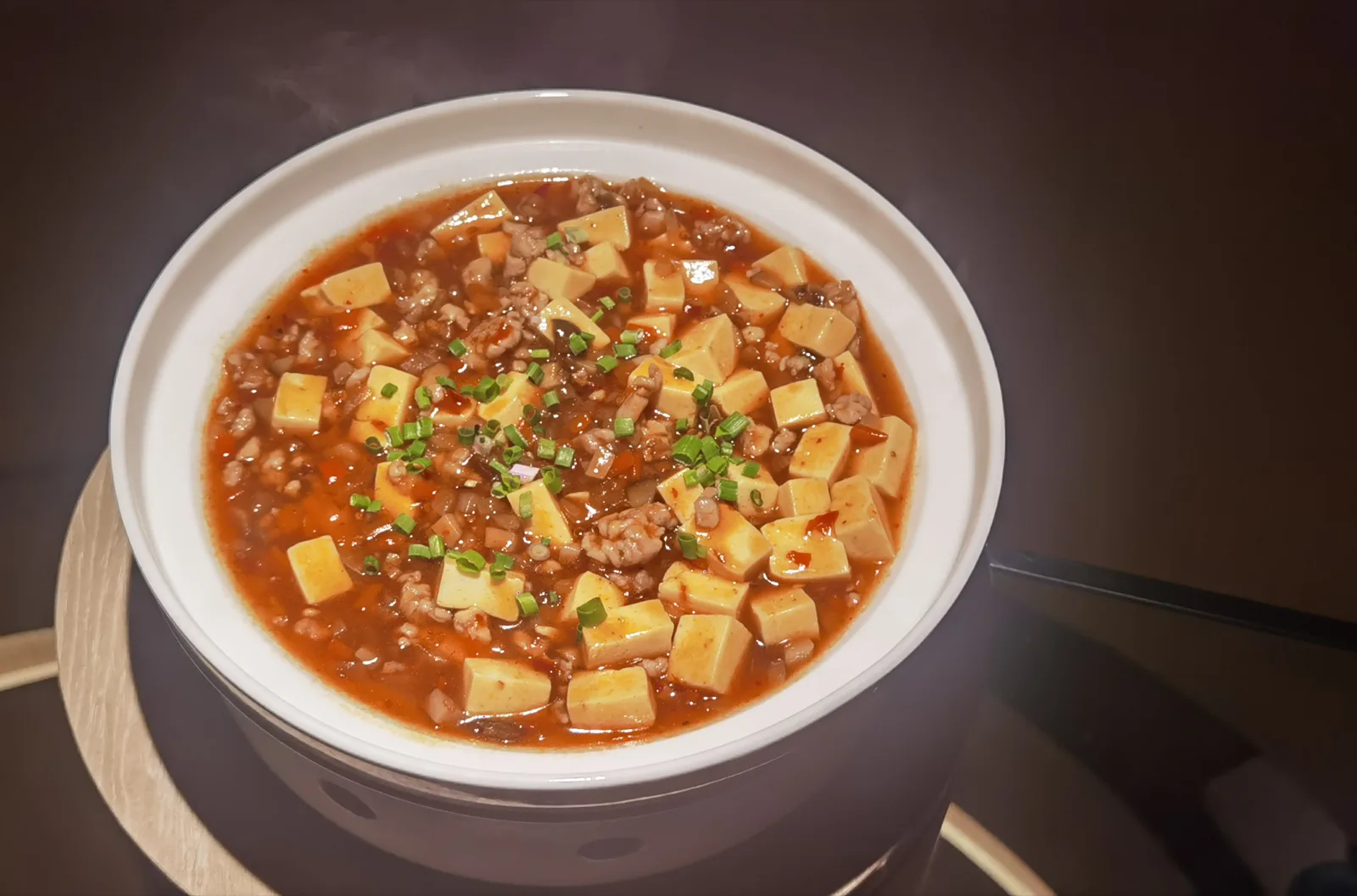 A bowl of mapo tofu set on a bamboo steamer tier, steam rising off the surface.